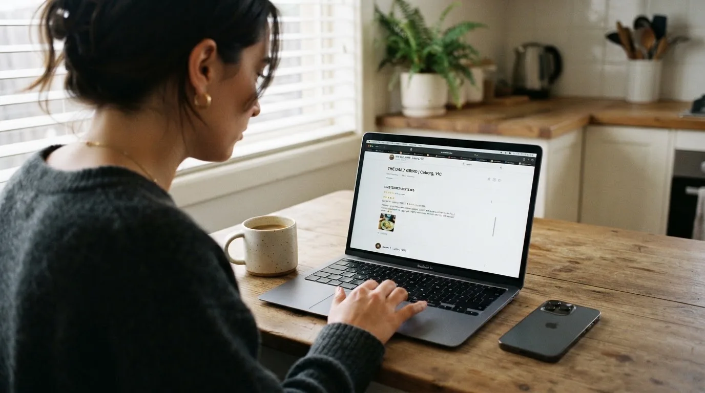 A person at a kitchen table scrolling through review pages on a laptop, phone face-down beside them.